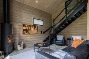 A modern living room with a wood-paneled wall, a black fireplace, a sofa with cushions, a coffee table, and a staircase. - Villa SnowLife B by Holiday in Lapland (Levi)