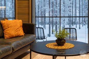 A modern interior with a leather sofa, a round table with a plant, and a view of a snowy forest through large windows. - Villa SnowLife B by Holiday in Lapland (Levi)