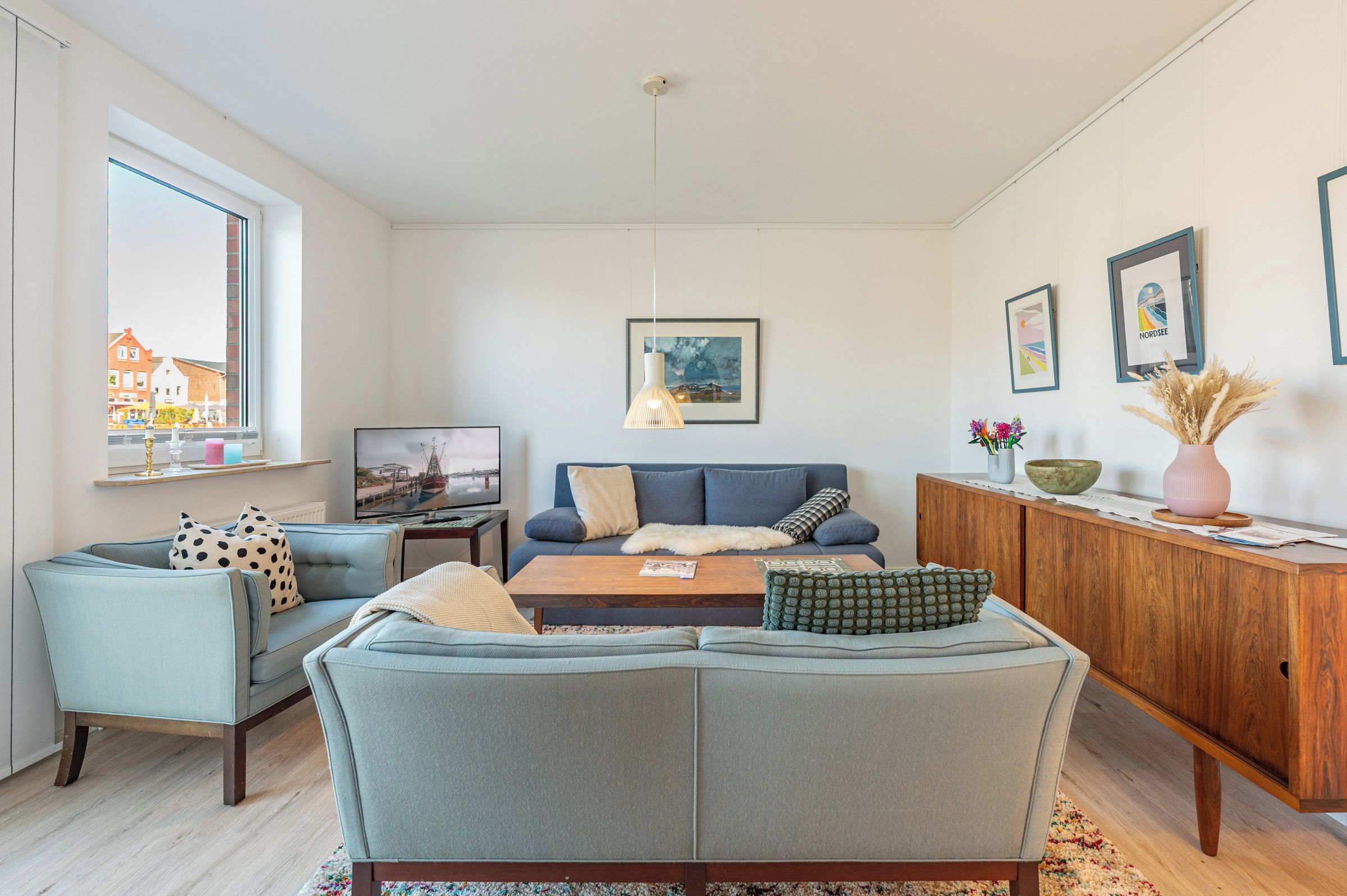 A modern living room with a sofa, armchairs, a coffee table, and a TV. There are framed pictures on the wall and a window with a view of buildings.