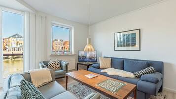 Een moderne woonkamer met een bank, een salontafel en een televisie. Er is een raam met uitzicht op gebouwen en een lamp.