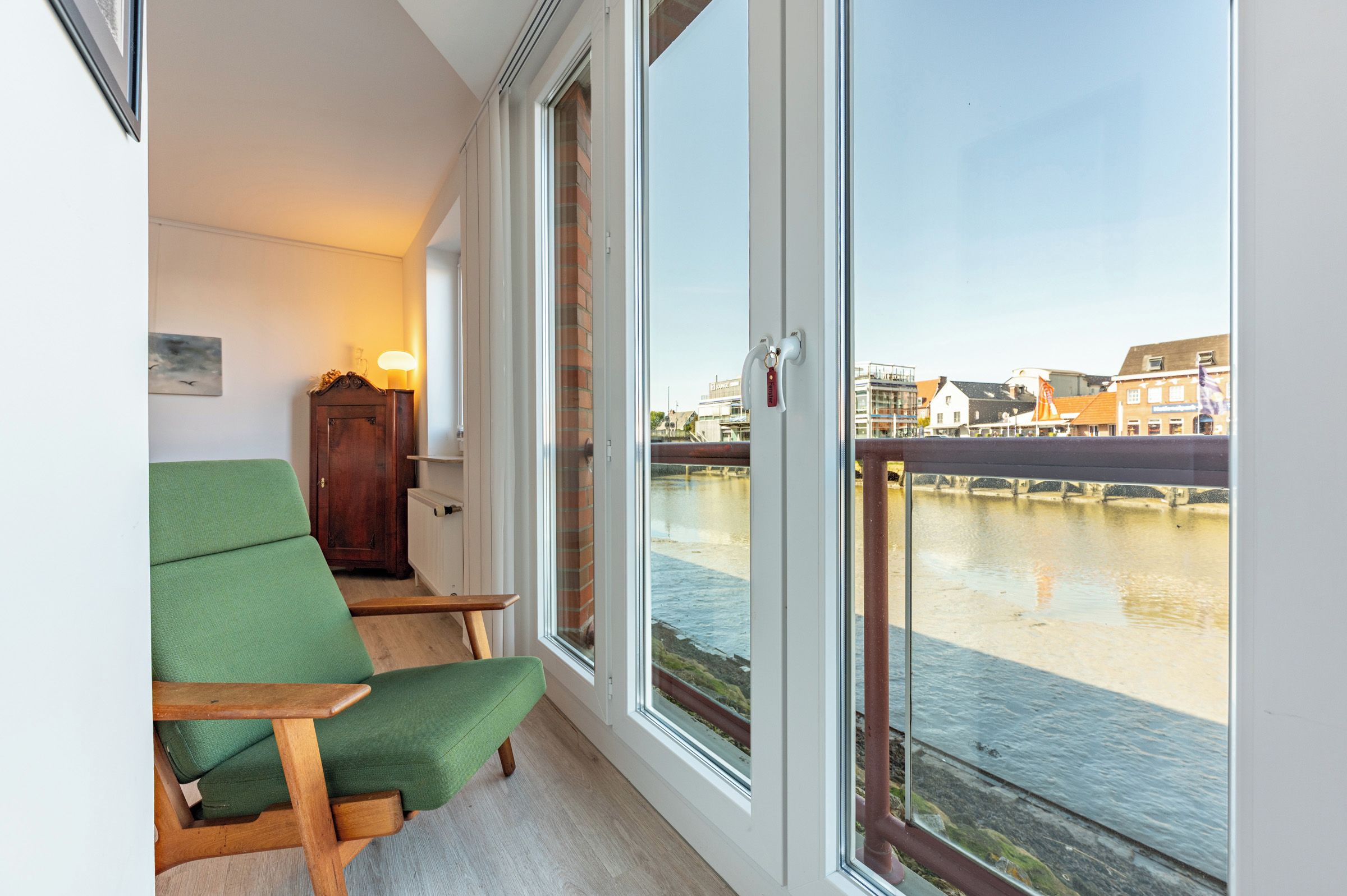 Ein Zimmer mit grünem Sessel, Holzschrank und Fenster mit Blick auf einen Fluss und Gebäude.