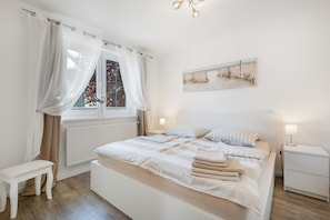 Ein Schlafzimmer mit einem Bett, Nachttischen, einem Fenster mit Vorhängen, einem Heizkörper und einem Bild an der Wand.