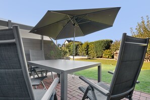 Eine Außenpatio mit Tisch und Stühlen, einem großen Sonnenschirm und Blick auf einen Garten sowie ein Haus im Hintergrund.