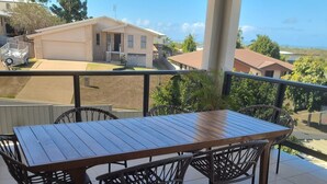 Eine überdachte Terrasse mit Holztisch und Korbstühlen, mit Blick auf ein Wohngebiet mit Häusern und blauem Himmel.