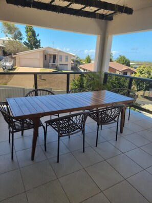 Un patio techado con mesa de madera y sillas de mimbre, con vista a un barrio residencial con casas y cielo despejado.