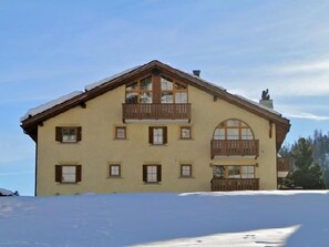 A two-story building with a balcony on the upper floor, large windows, and a snow-covered ground. - Two-room apartment on the top floor (4th floor), approximately 60 m². (Celerina/Schlarigna)