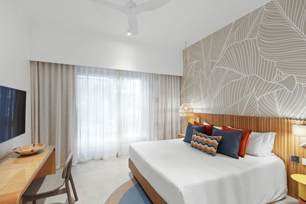 A hotel room with a bed, a desk, a television, and a large window with curtains.