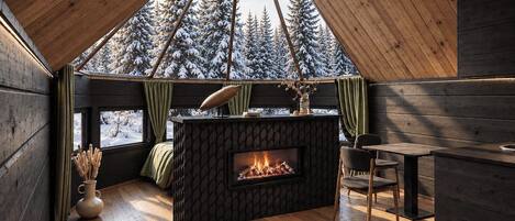 Interior acogedor de una cabaña con techo de madera, chimenea, comedor y vista a árboles nevados.