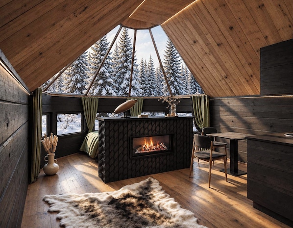 Interior acogedor de una cabaña con techo de madera, chimenea, comedor y vista a árboles nevados.