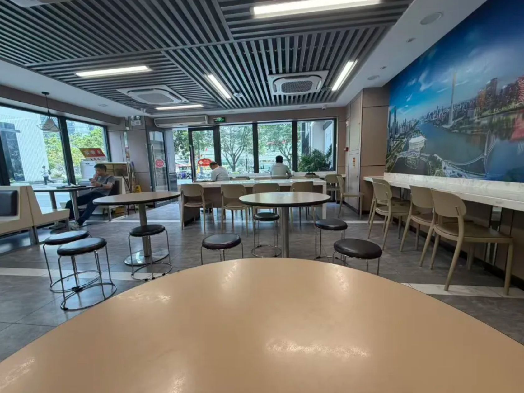 A modern indoor dining area with tables and chairs, large windows, and a cityscape mural on the wall.