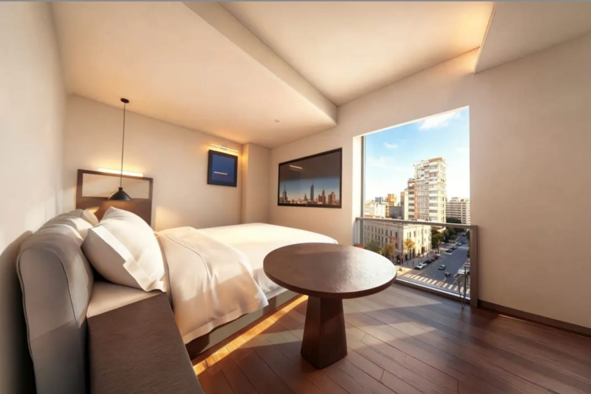 A modern hotel room with a bed, a sofa, a round table, and a city view through a large window.