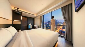 A hotel room with a large bed, a desk, a chair, a small table with a vase, and a view of a cityscape through a large window. - Ruilan (Guangzhou)