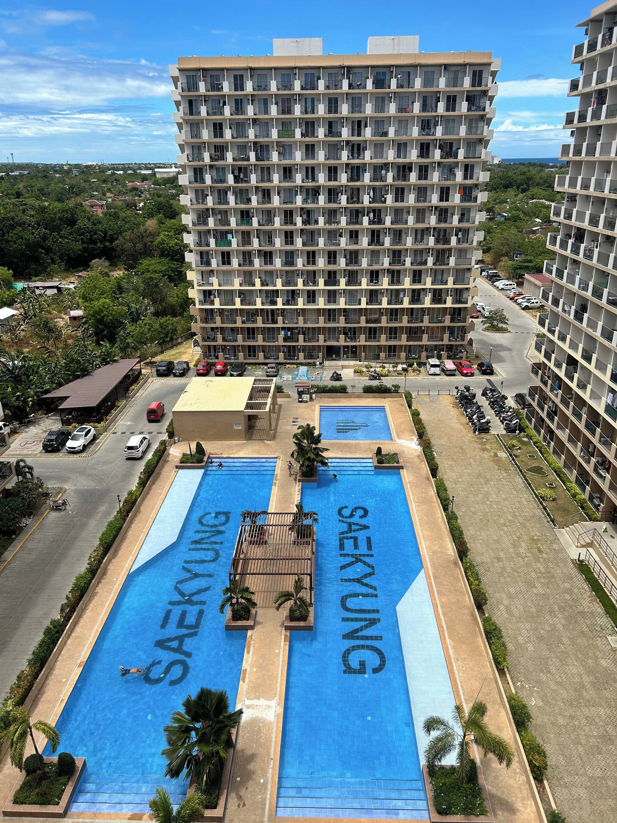 A multi-story building with a rooftop pool area labeled 'SAEKYU' and a parking lot with several cars.