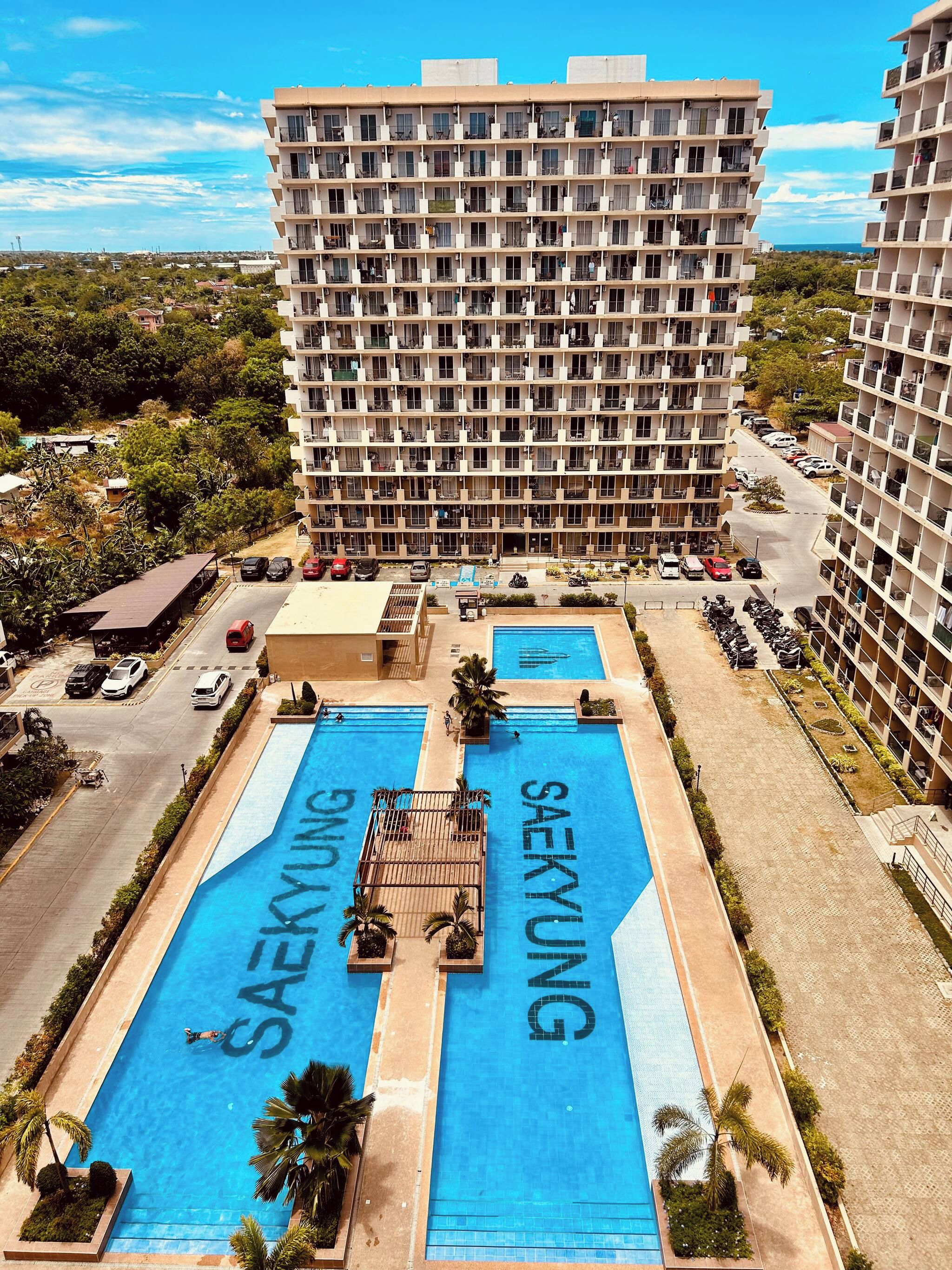 Hotel de varios pisos con piscina en la azotea, rodeado de coches aparcados y vegetación.