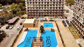 Hotel de varios pisos con piscina en la azotea, rodeado de coches aparcados y vegetación.