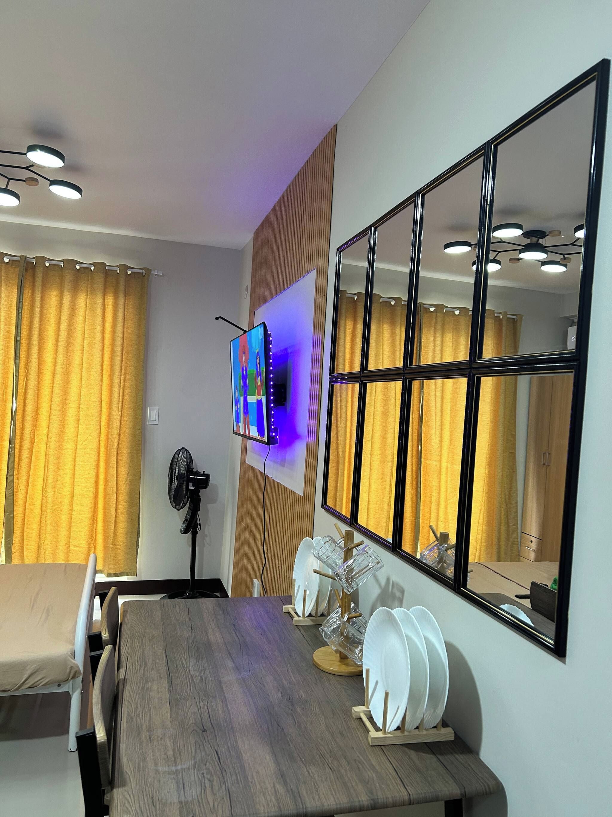 A dining area with a wooden table, chairs, a wall-mounted TV, and a large mirror reflecting the room.