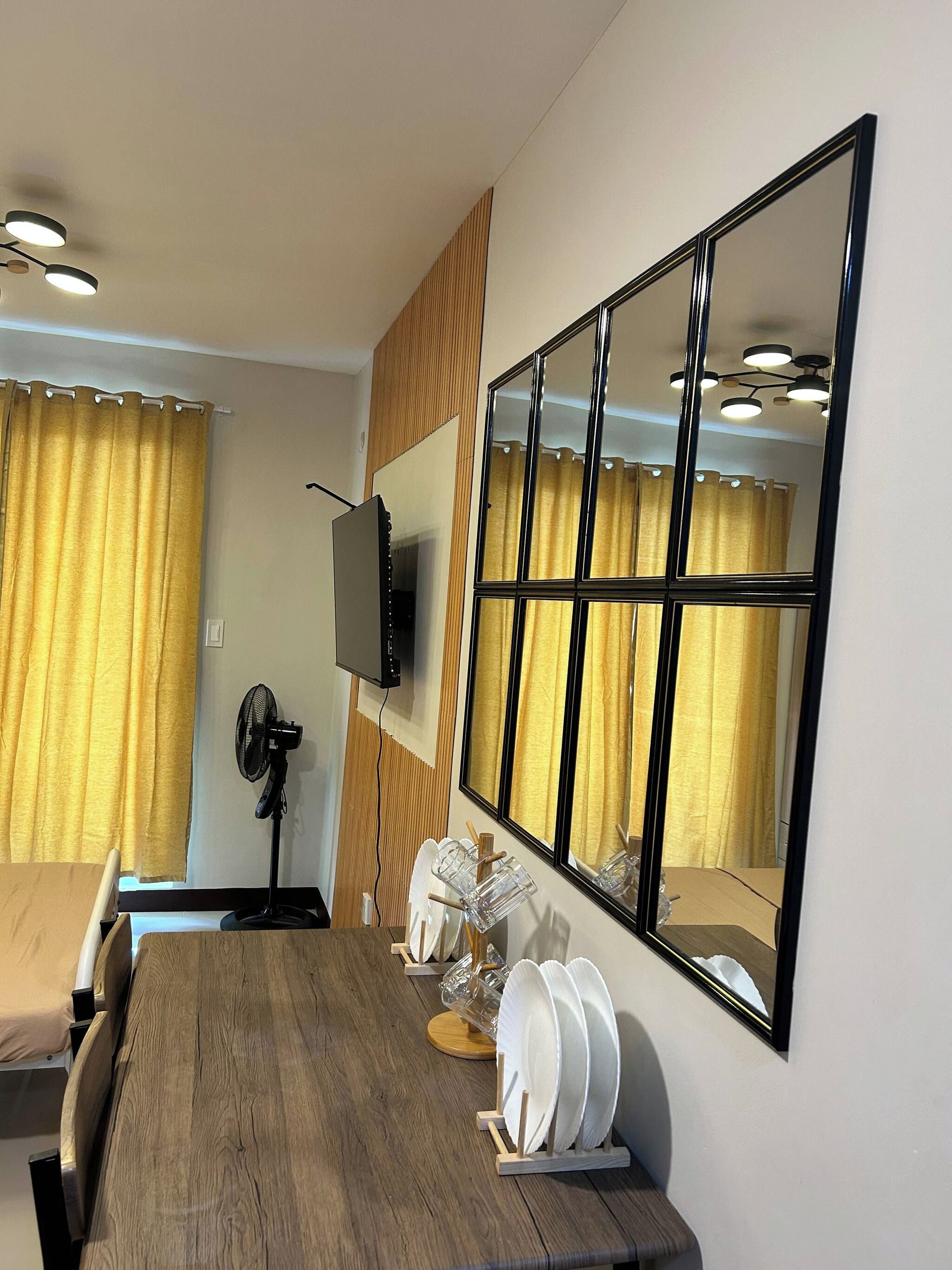 Una habitación de hotel con una mesa de comedor de madera, un televisor de pantalla plana montado en la pared, un ventilador de pie y un espejo grande que refleja la habitación.