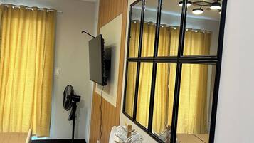 Una habitación de hotel con una mesa de comedor de madera, un televisor de pantalla plana montado en la pared, un ventilador de pie y un espejo grande que refleja la habitación.