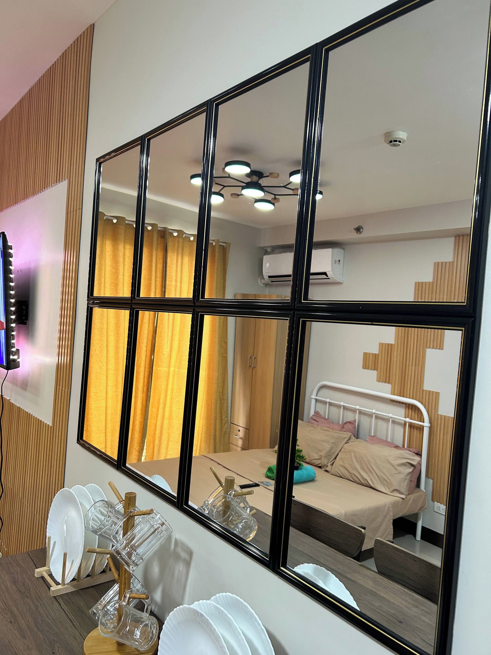 Une chambre d’hôtel avec un grand miroir, un lit, une table de chevet avec une lampe, un climatiseur fixé au mur et une fenêtre avec des rideaux.