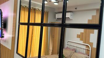 Une chambre d’hôtel avec un grand miroir, un lit, une table de chevet avec une lampe, un climatiseur fixé au mur et une fenêtre avec des rideaux.