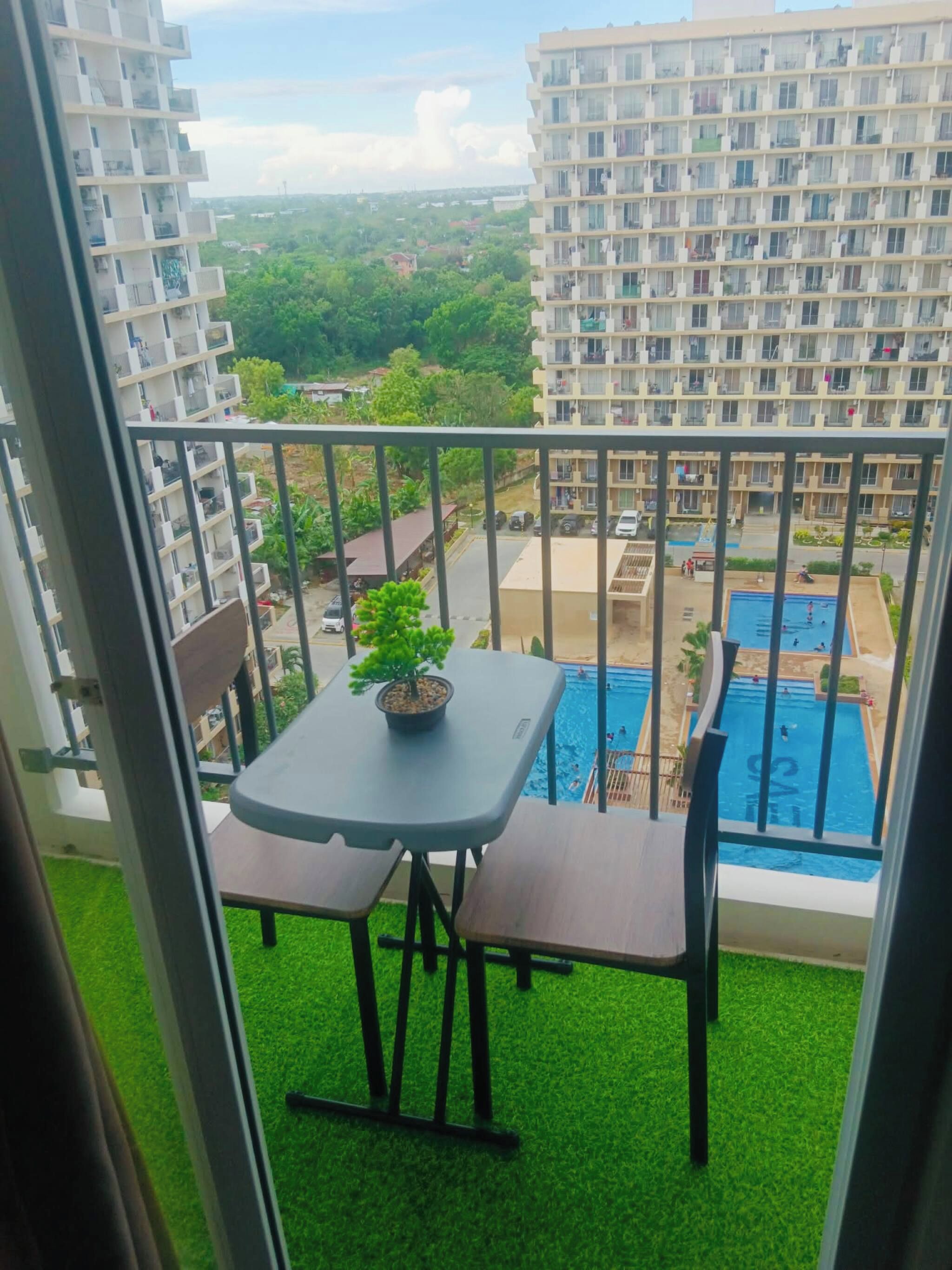 Un balcón con una mesa y sillas, una planta pequeña en maceta y vistas a una piscina y edificios altos.