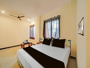 A hotel room with a bed, a bedside table, a telephone, a window with curtains, a ceiling fan, and artwork on the wall.