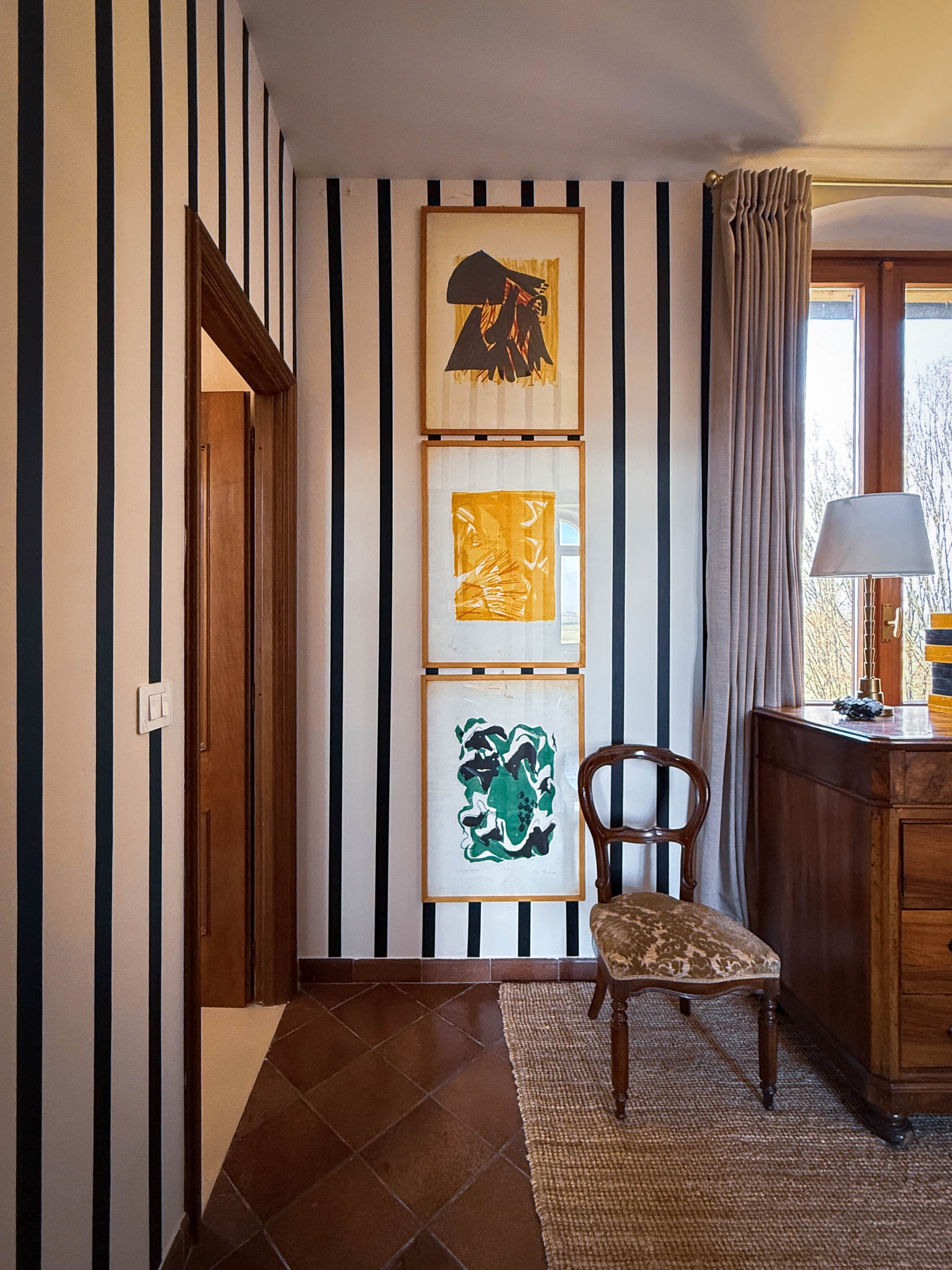 Un corridoio con carta da parati a righe, una porta in legno, una sedia, una scrivania con una lampada e opere d'arte incorniciate al muro.