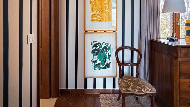 Un corridoio con carta da parati a righe, una porta in legno, una sedia, una scrivania con una lampada e opere d'arte incorniciate al muro.