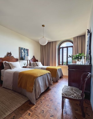 A hotel room with two beds, a desk, a chair, a large window with curtains, a painting on the wall, and a hanging light fixture. - Le Nonne Suite (Chieri)