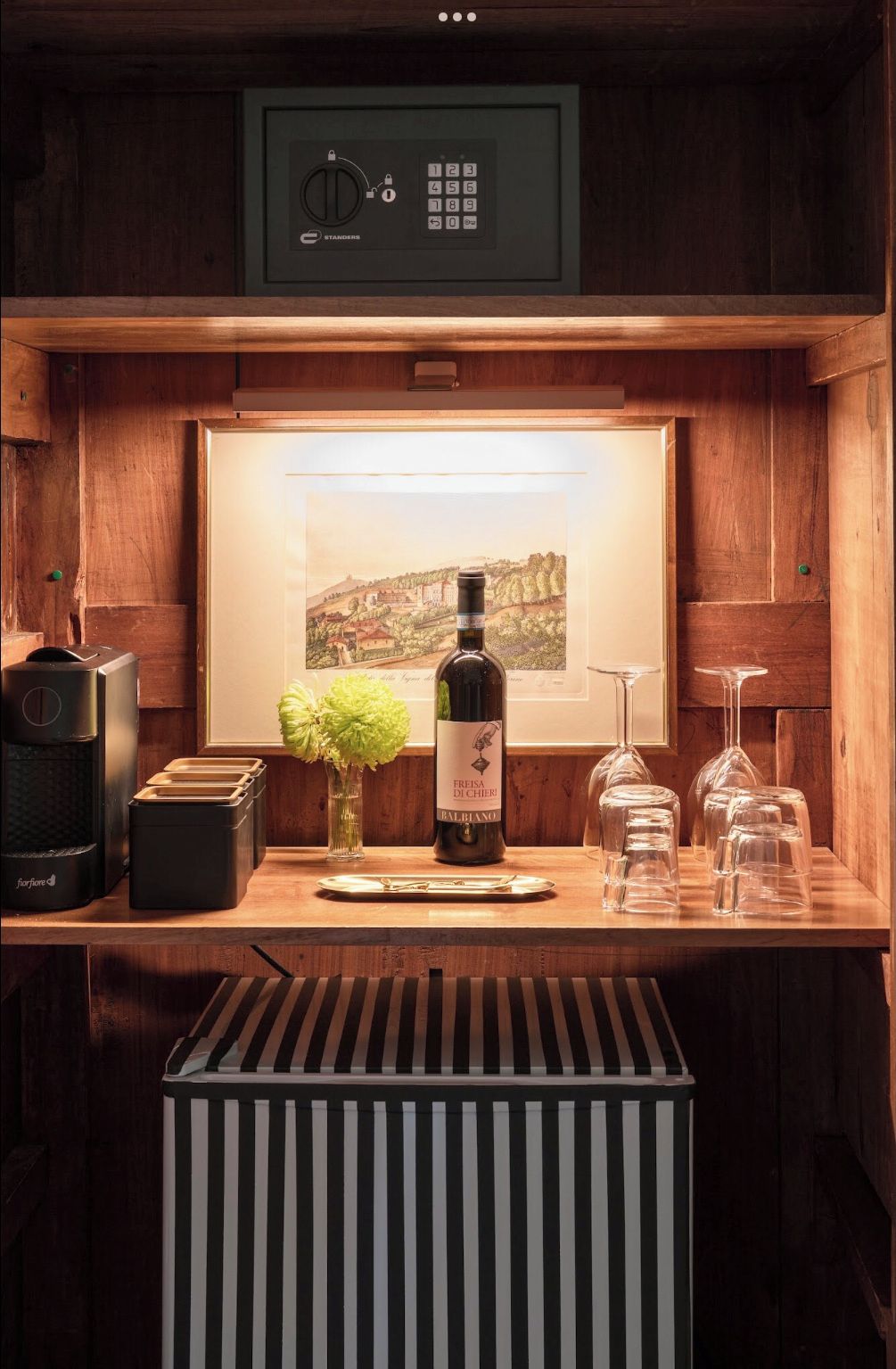 A wooden bar area with a framed picture, a bottle of wine, glasses, and a coffee machine.