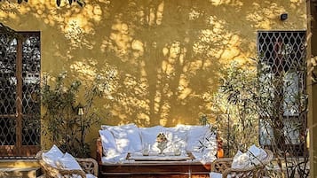 Área de estar ao ar livre banhada pelo sol, com um banco de madeira e cadeiras de vime, cercada por plantas e uma parede amarela.