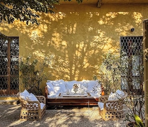 A sunlit outdoor seating area with a wooden bench and wicker chairs, surrounded by plants and a yellow wall.