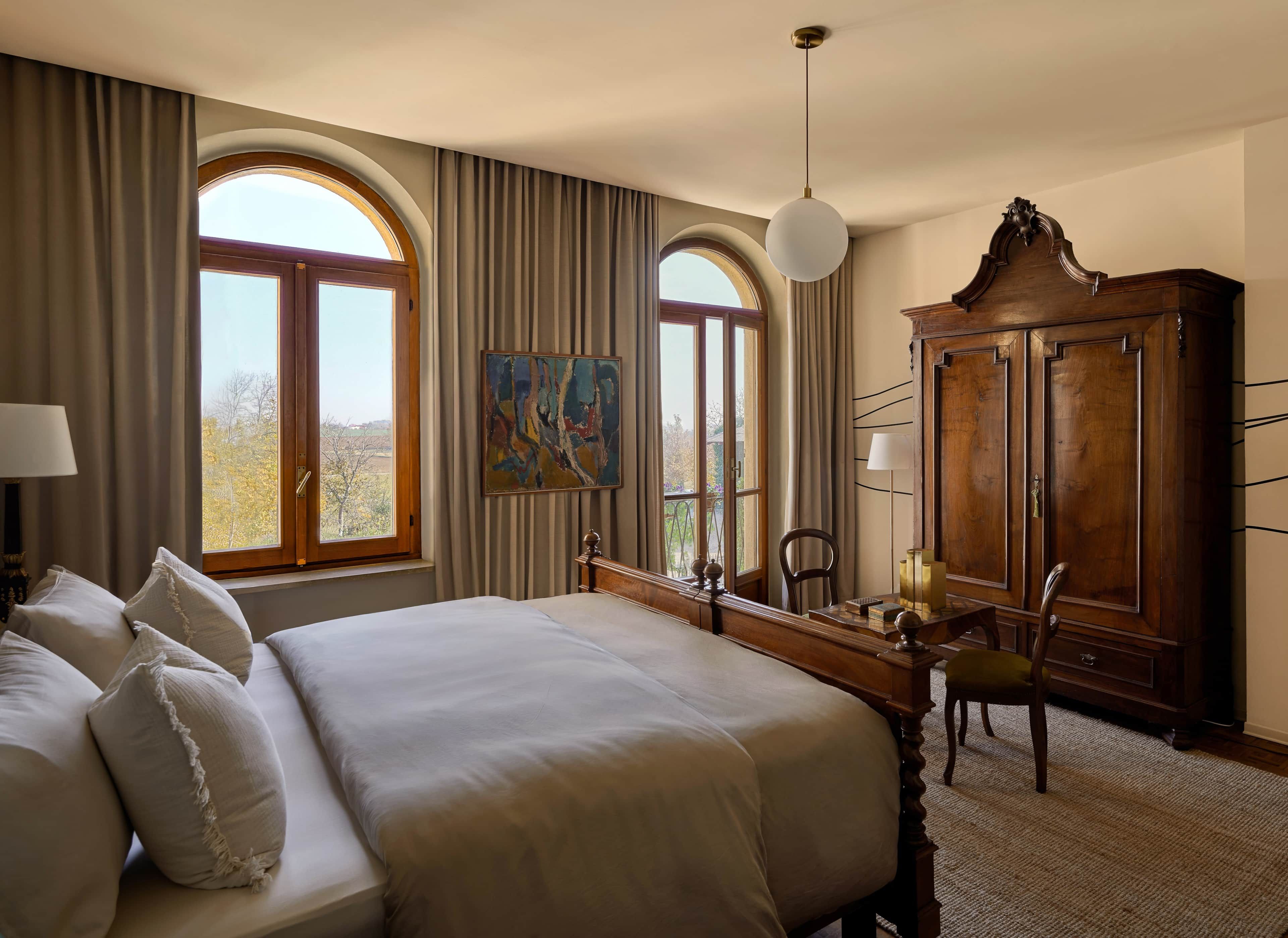 A hotel room with a large bed, a desk with a chair, a wardrobe, and two windows with curtains.