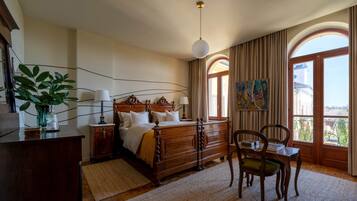 A bedroom with a wooden bed, a desk with chairs, a large window with curtains, and a painting on the wall.