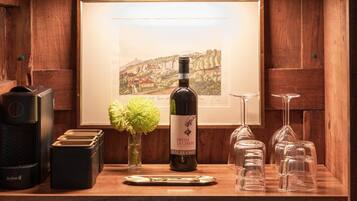 A wooden bar area with a safe, a framed landscape painting, a bottle of wine, a vase with flowers, and wine glasses.
