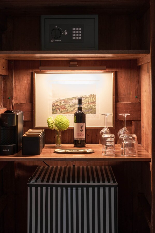 A wooden bar area with a safe, a framed landscape painting, a bottle of wine, a vase with flowers, and wine glasses.