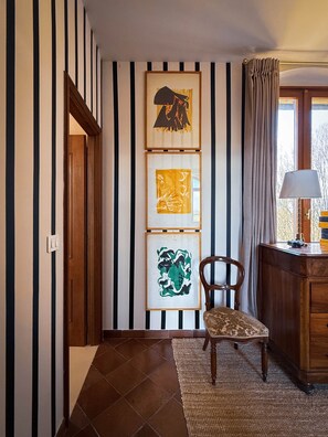 A hallway with striped wallpaper, a wooden door, a chair, a desk with a lamp, and framed artwork on the wall.