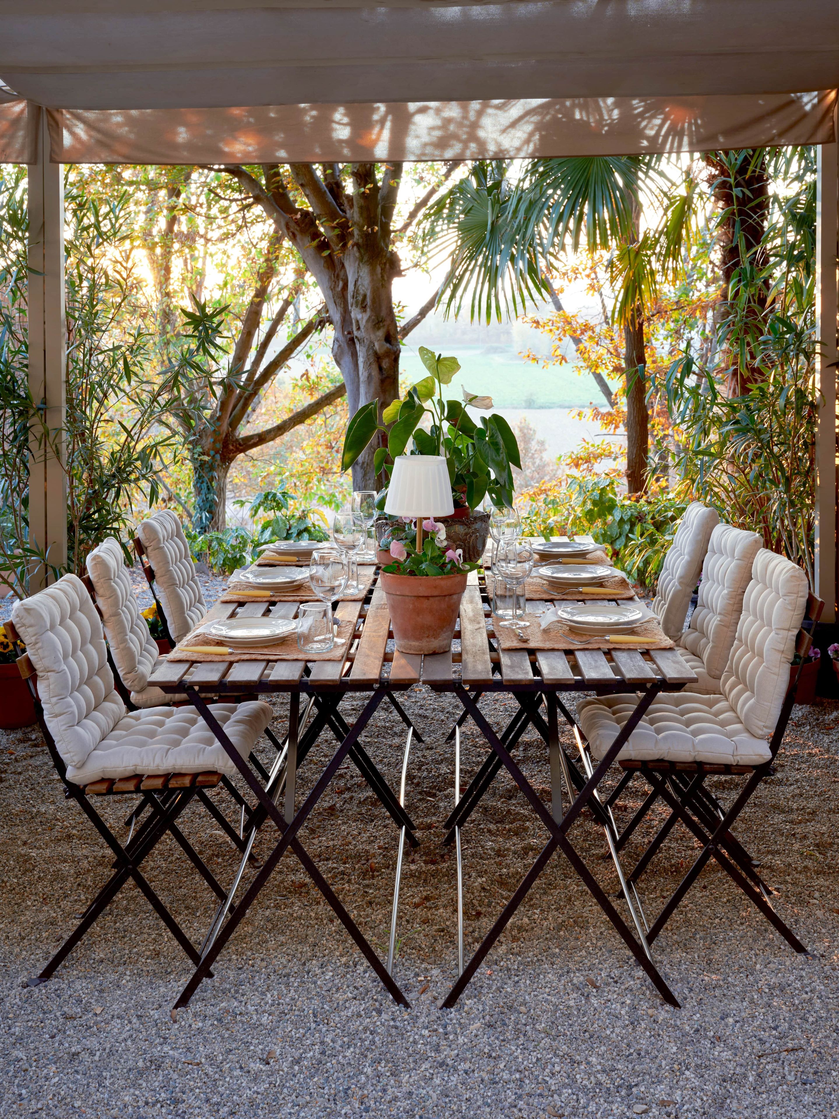 An outdoor dining area with a table set for four, surrounded by cushioned chairs, under a pergola with a view of trees and a body of water.
