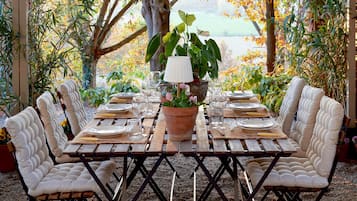 An outdoor dining area with a table set for four, surrounded by cushioned chairs, under a pergola with a view of trees and a body of water.