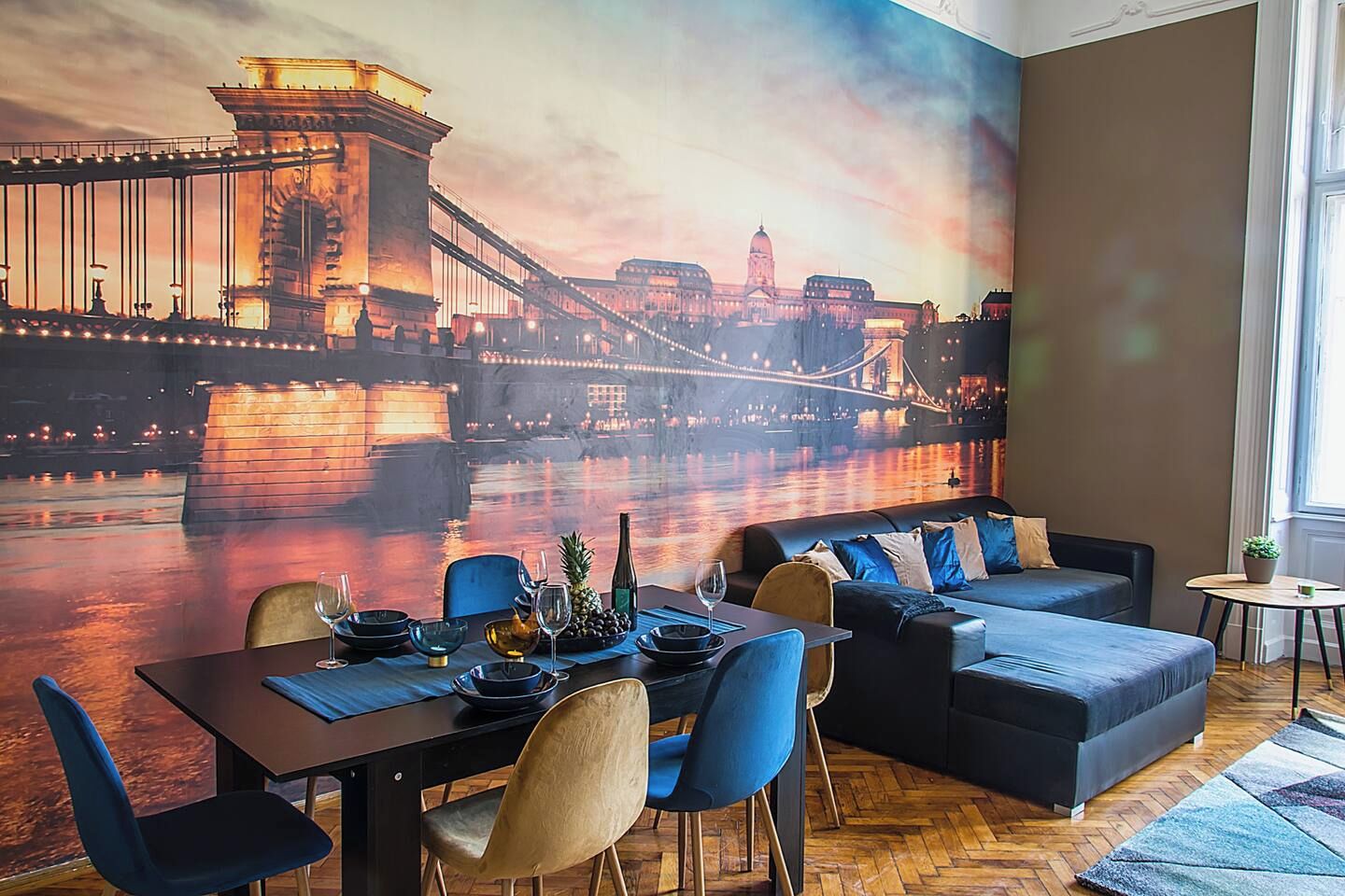 A dining area with a mural of a bridge, a table set for two, and a sofa.