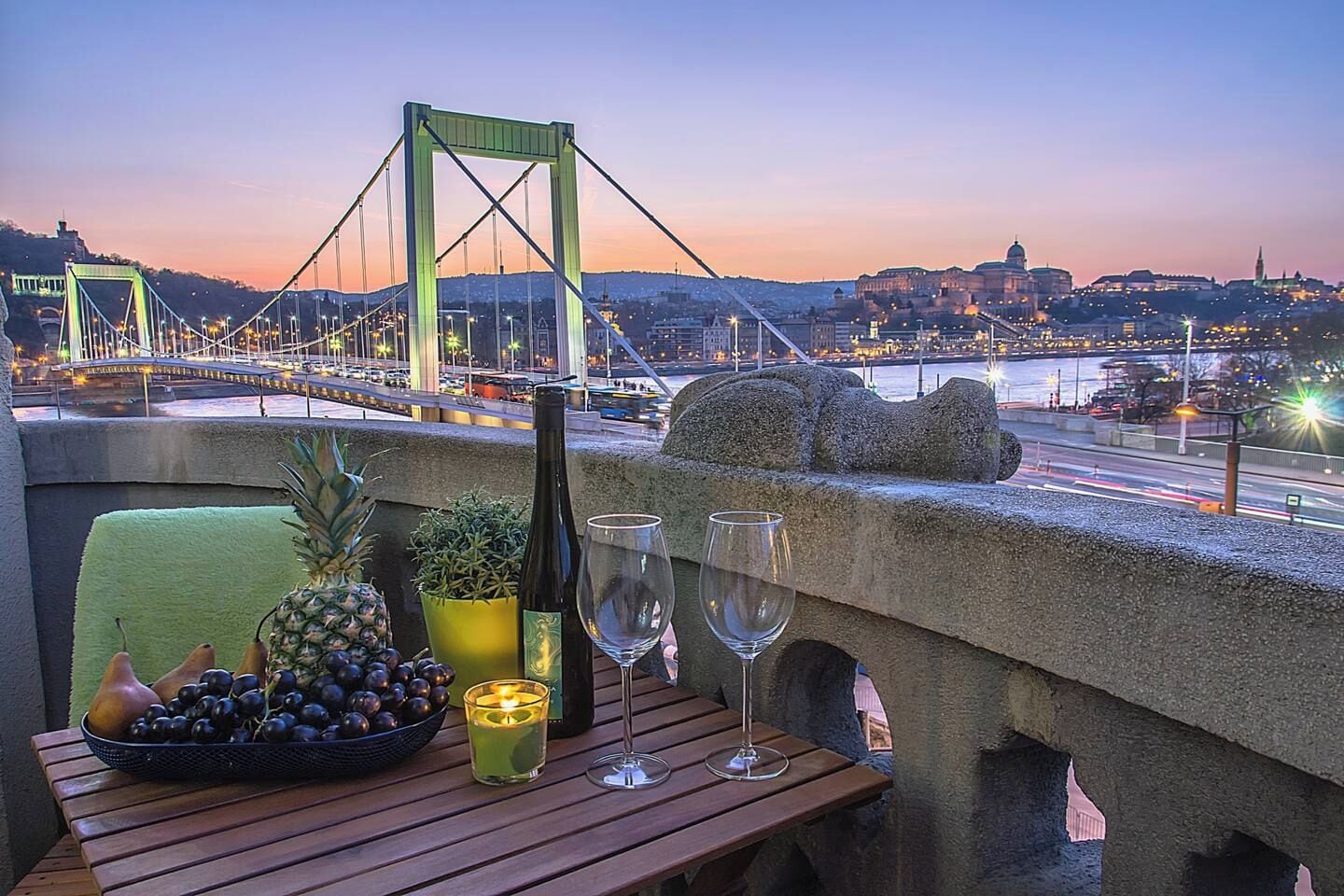 Une table avec une bouteille, des verres, des fruits et une bougie, donnant sur un paysage urbain avec un pont et des bâtiments.