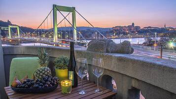 Une table avec une bouteille, des verres, des fruits et une bougie, donnant sur un paysage urbain avec un pont et des bâtiments.