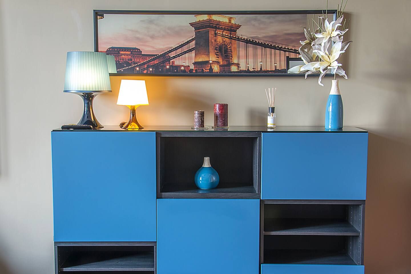 A blue cabinet with a lamp, a vase, and a framed picture of a bridge.