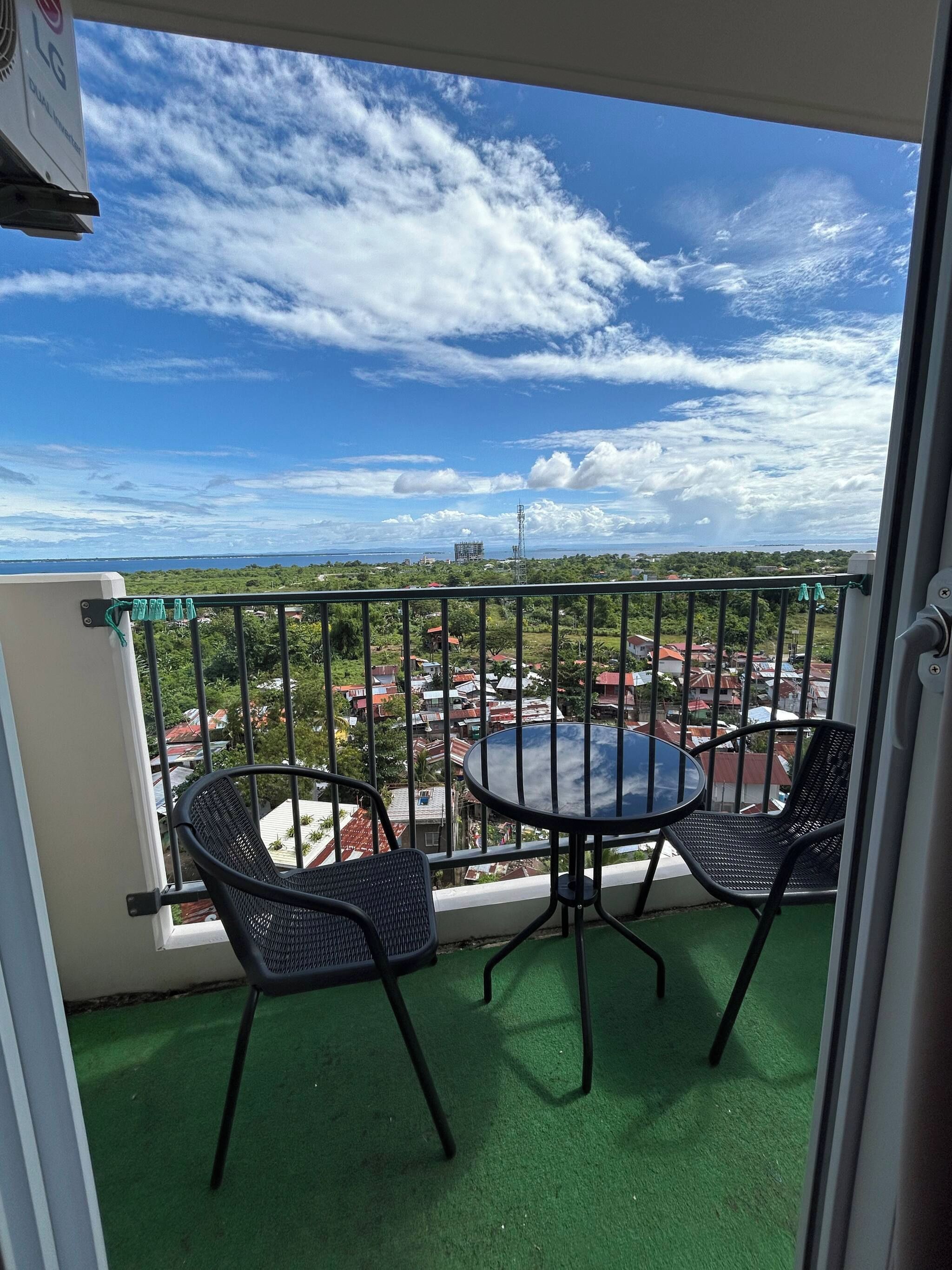 A balcony with two chairs and a table, overlooking a cityscape with buildings and greenery.