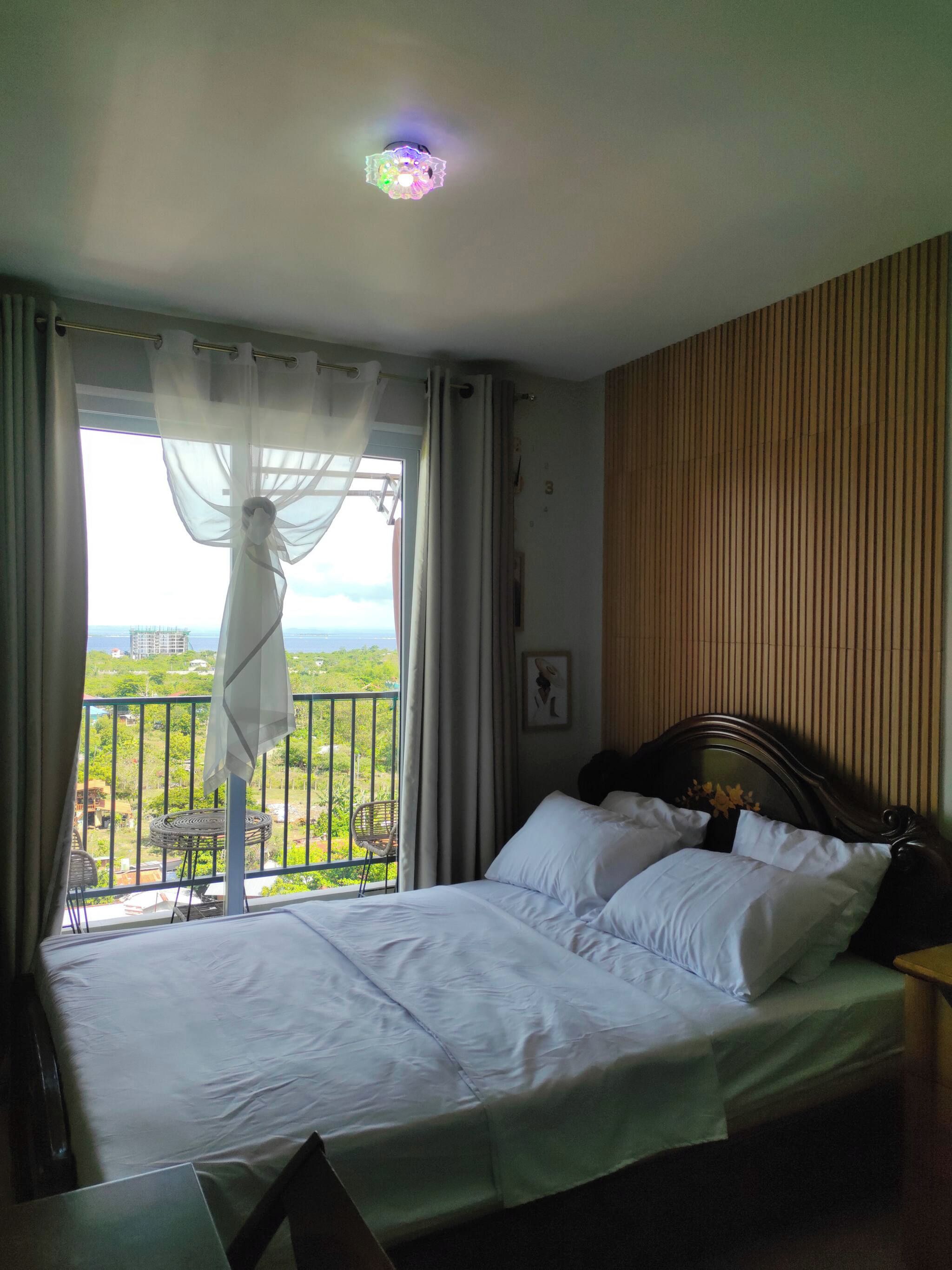 Habitación de hotel con cama, vista a zonas verdes y edificios, y balcón con una silla.