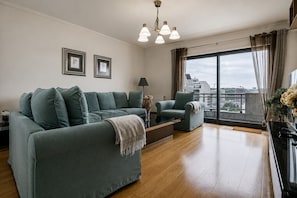 Ein modernes Wohnzimmer mit einem Ecksofa, einem zusätzlichen Sofa, einem Couchtisch und einem Beistelltisch. Ein großes Fenster bietet Blick auf Gebäude und einen Balkon.