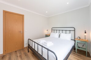 A bedroom with a wooden door, a metal bed frame, white bedding, two bedside tables with lamps, and a clock on one table. - Nomad's Family - 2-Bedroom Apartment at The Docks in Porto (Porto)