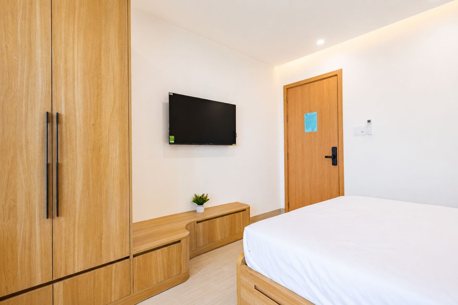 A bedroom with a bed, wooden wardrobe, a small plant, a flat-screen TV, and a door with a lock.