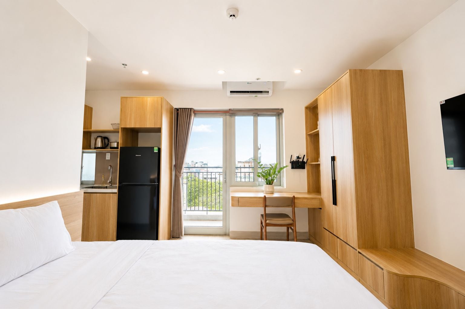 Habitación de hotel con cama, escritorio, refrigerador, planta en maceta y ventana con vista a la ciudad.