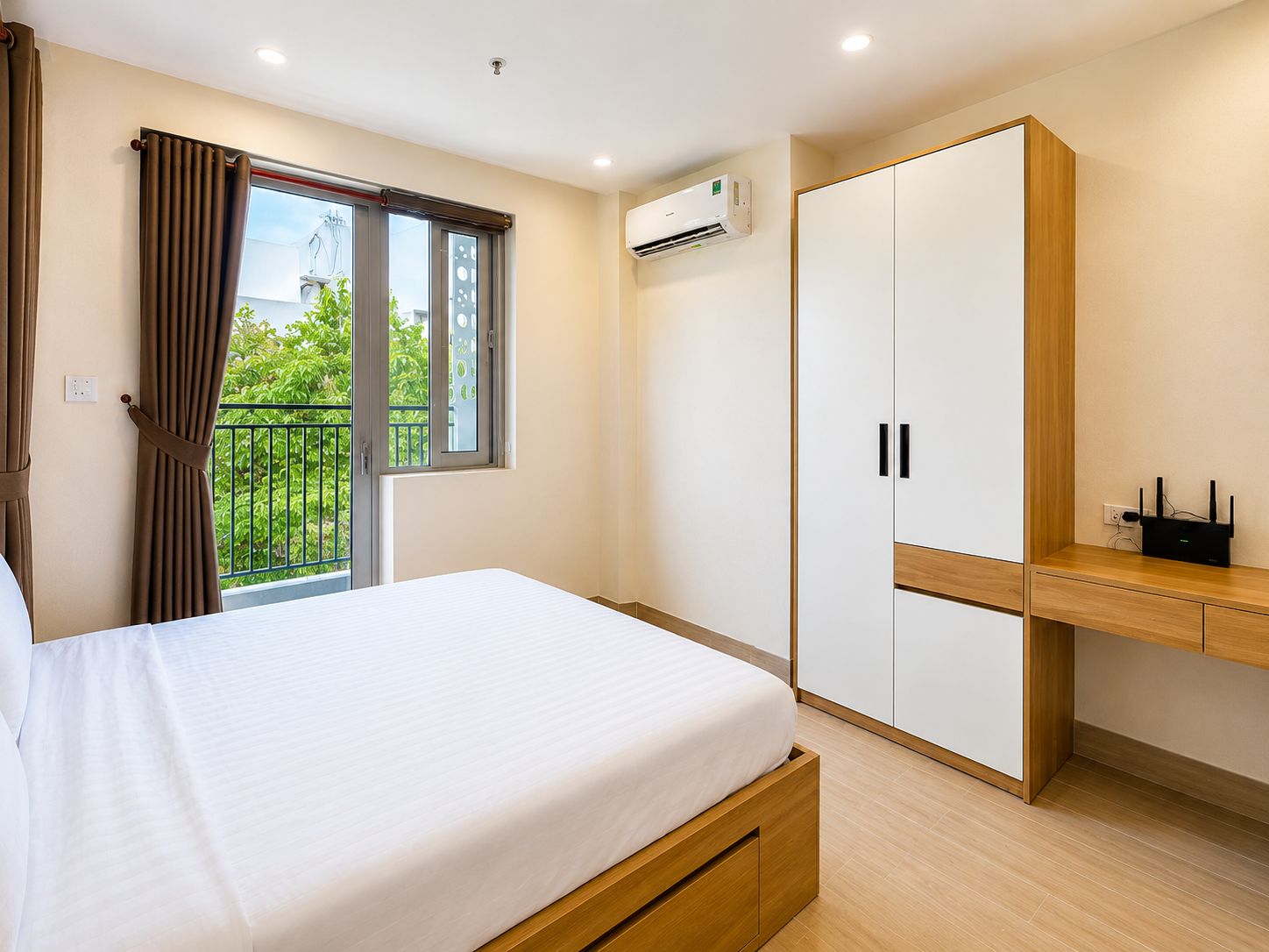 A bedroom with a bed, a wardrobe, a desk, an air conditioning unit, and a view of greenery through a window.